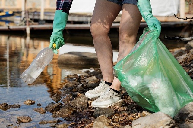 Foto van dame met handschoenen in korte broek die zwerfvuil opraapt aan de waterkant en die in een plastic afvalzak stopt.