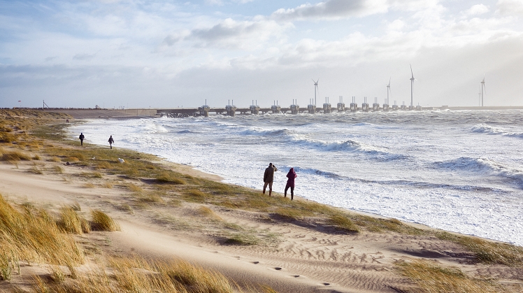 Foto van stormachtige kust aan de Oosterschelde, wandelende mensen aan de kust en de Oosterscheldekering op de achtergrond.