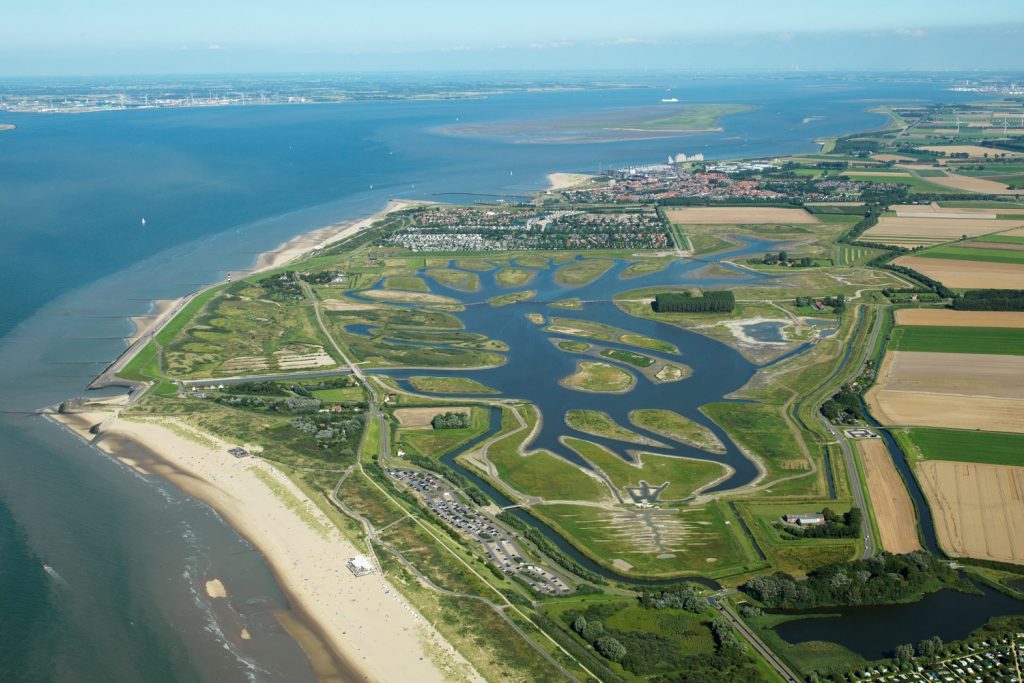 Luchtfoto van het natuur- en recreatiegebied Waterdunen. Langs de kust zie je duinen, groene vegetatie, plassen en polders.