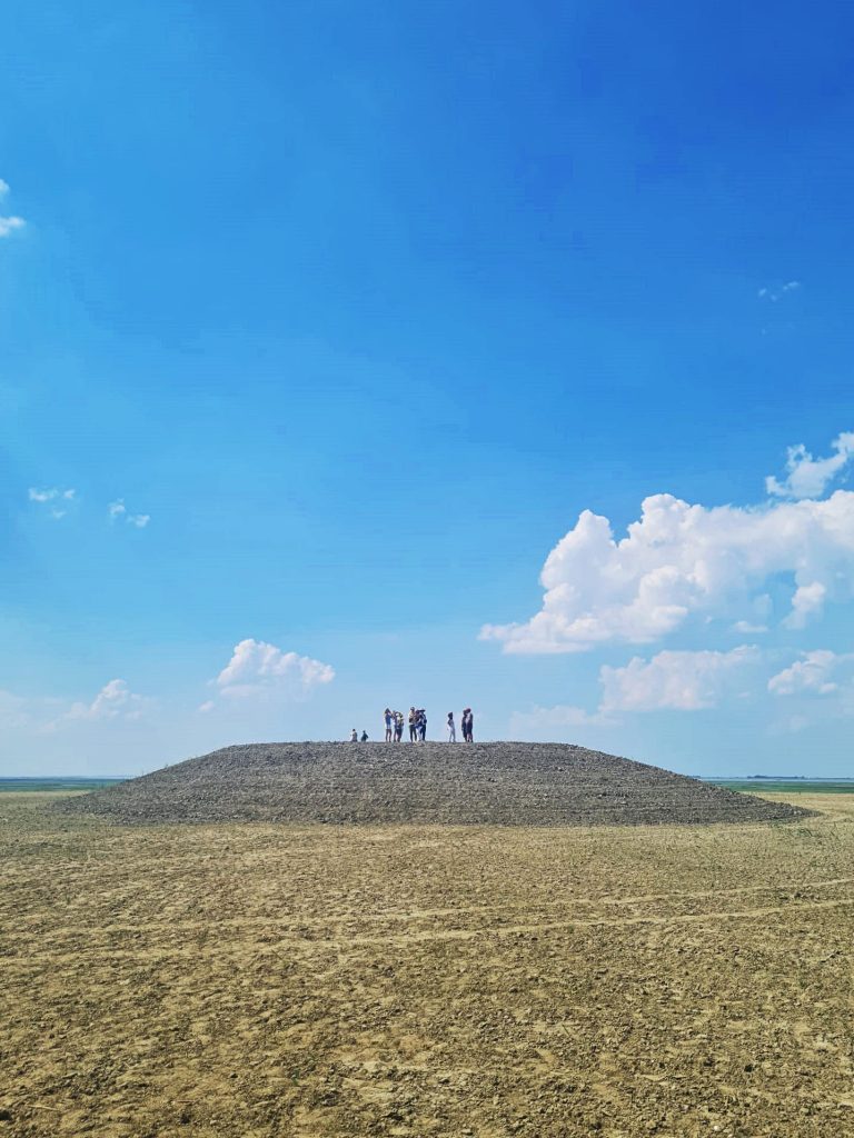 Een heuvel met vlakke top in het landschap. Op de top staat een groep mensen. De lucht is blauw met enkele wolken.