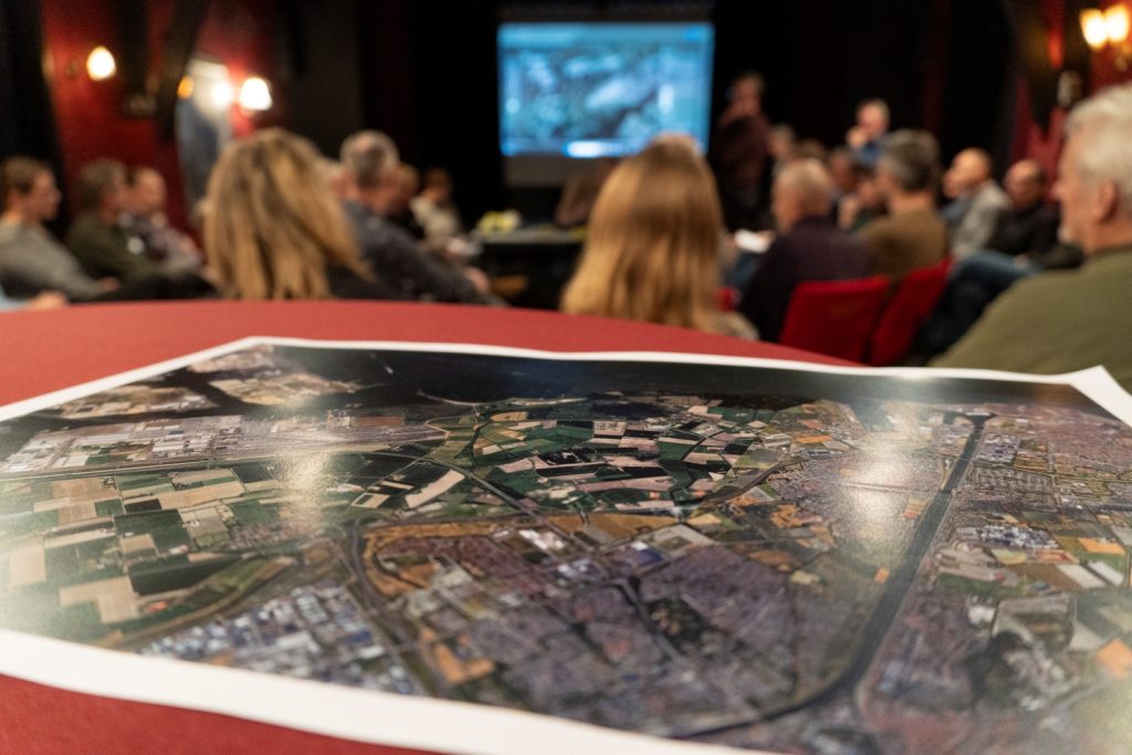 Close-upfoto van een afgedrukte plattegrond. De afdruk ligt op een rode tafel. In de achtergrond zijn een groep mensen wazig in beeld.