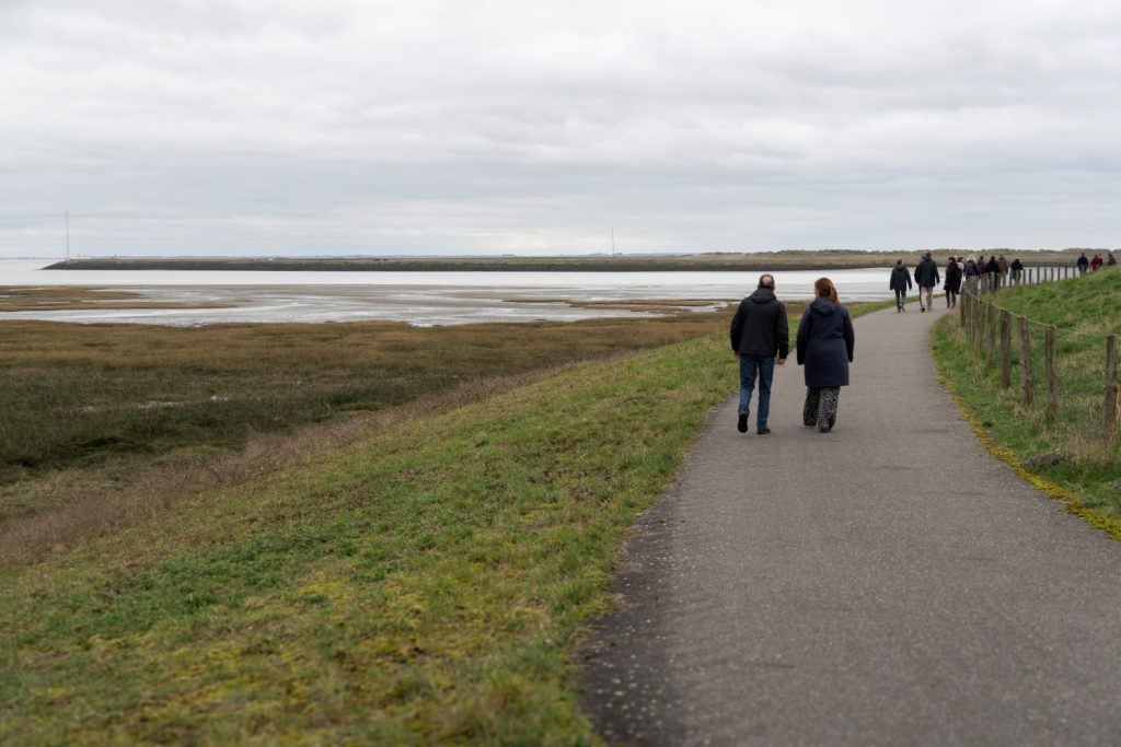 Een groep mensen loopt over een geasfalteerde dijk. De rechterkant is afgezet, aan de linkerkant is water en lage begroeiing zichtbaar.