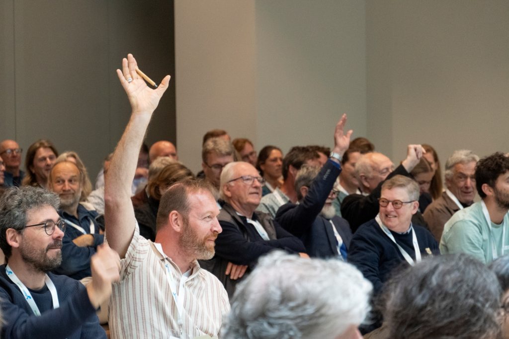 Een zaal vol mensen. Scherp in beeld is een man die zijn hand opsteekt.