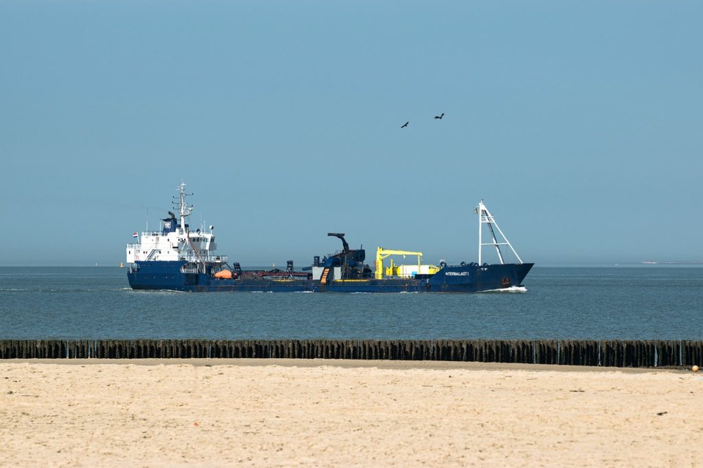 Een baggerschip op zee, met in de voorgrond zand en een rij paaltjes.