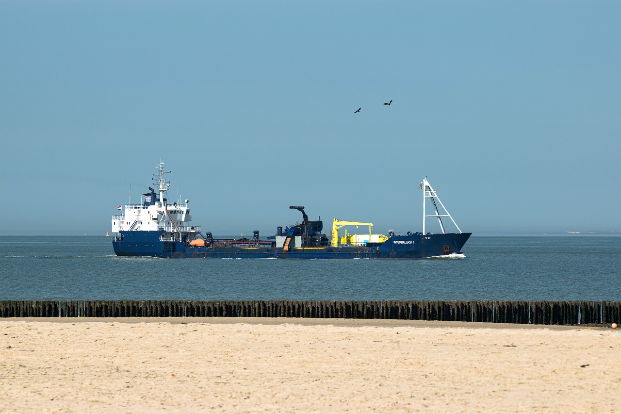 Een baggerschip op zee, met in de voorgrond zand en een rij paaltjes.