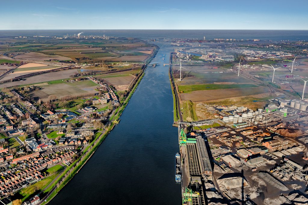 Luchtfoto van het kanaal Gent-Terneuzen. In de verte is de Westerschelde te zien. Aan beide zijden van het kanaal zijn woningen, industrie en landbouw te zien.