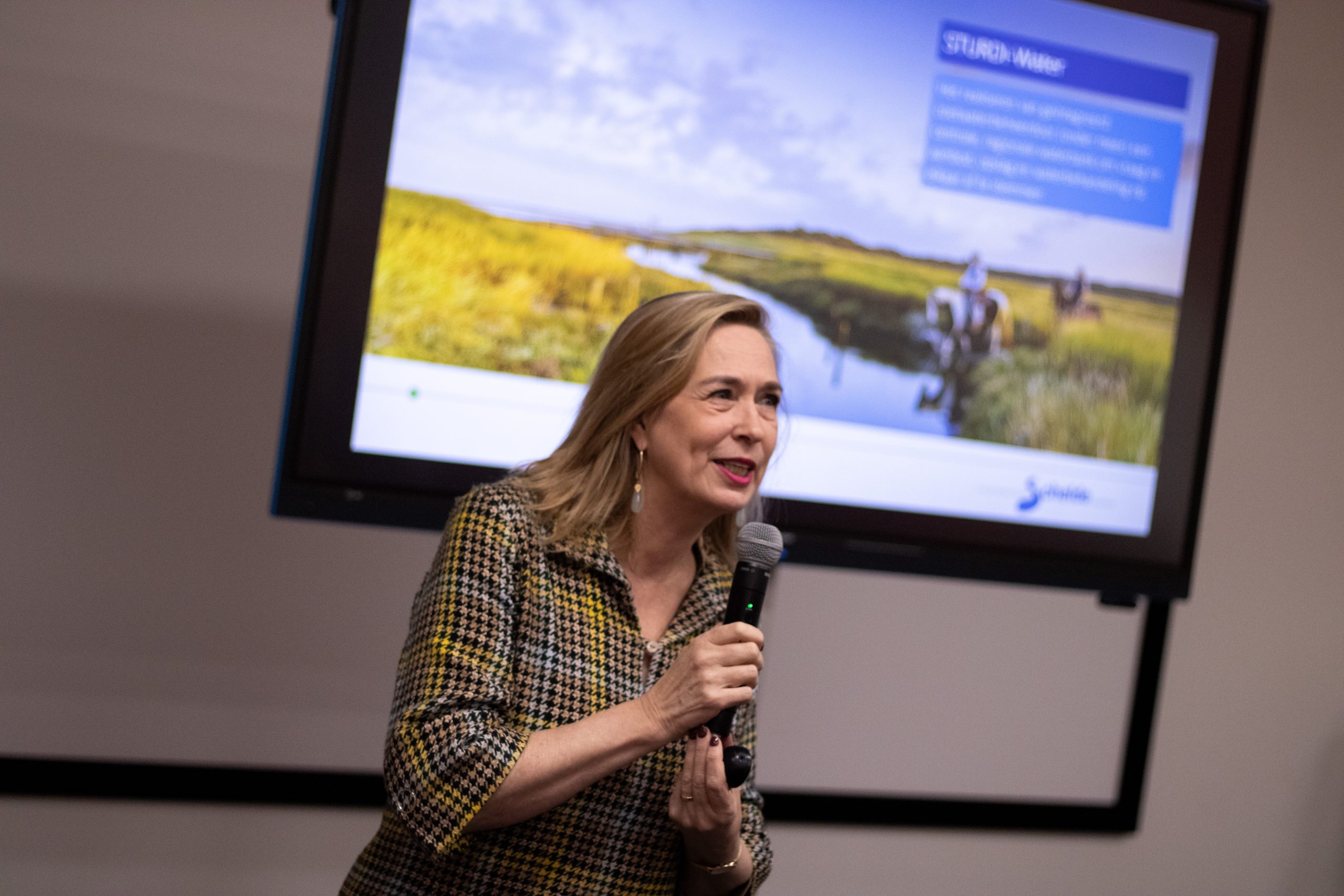 Een foto van voorzitter Barbara Oomen. Een vrouw met blond haar en een subtiel geblokte jurk. Ze houdt een microfoon vast. Op de achtergrond is vaag de presentatie van het symposium te zien.