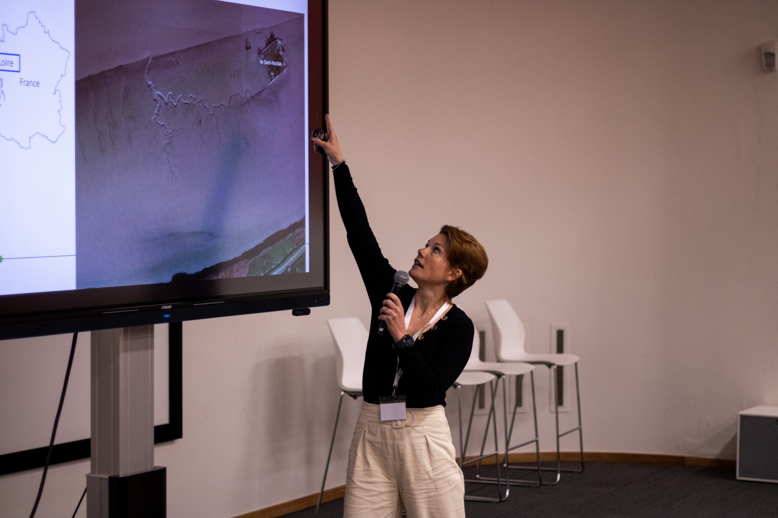 Foto van Jill Hansen. Een jonge vrouw met kort, bruin haar. Ze wijst naar haar presentatie en houdt een microfoon vast.