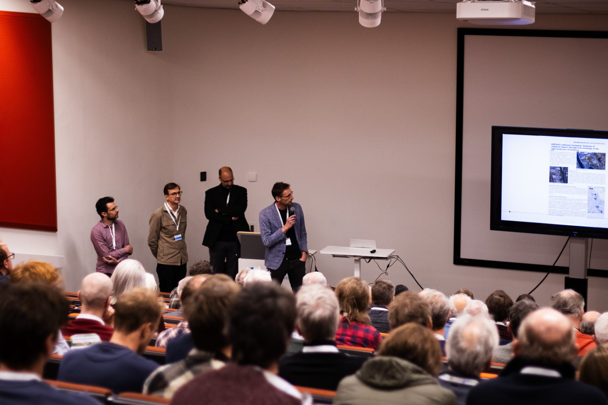 Foto van de achterhoofden van het publiek. We zien de vier presentatoren van de poster pitches van veraf. Eén van hen presenteert en houdt een microfoon vast.