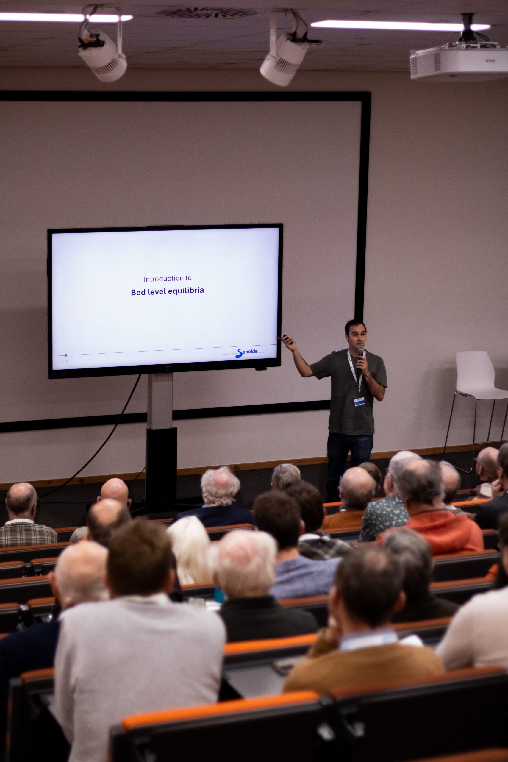 Foto van de achterhoofden van het publiek. In de verte zie je Greg Fivash wijzen naar zijn presentatie, met in zijn andere hand een microfoon. Hij heeft zwart, kort haar en een groene trui.