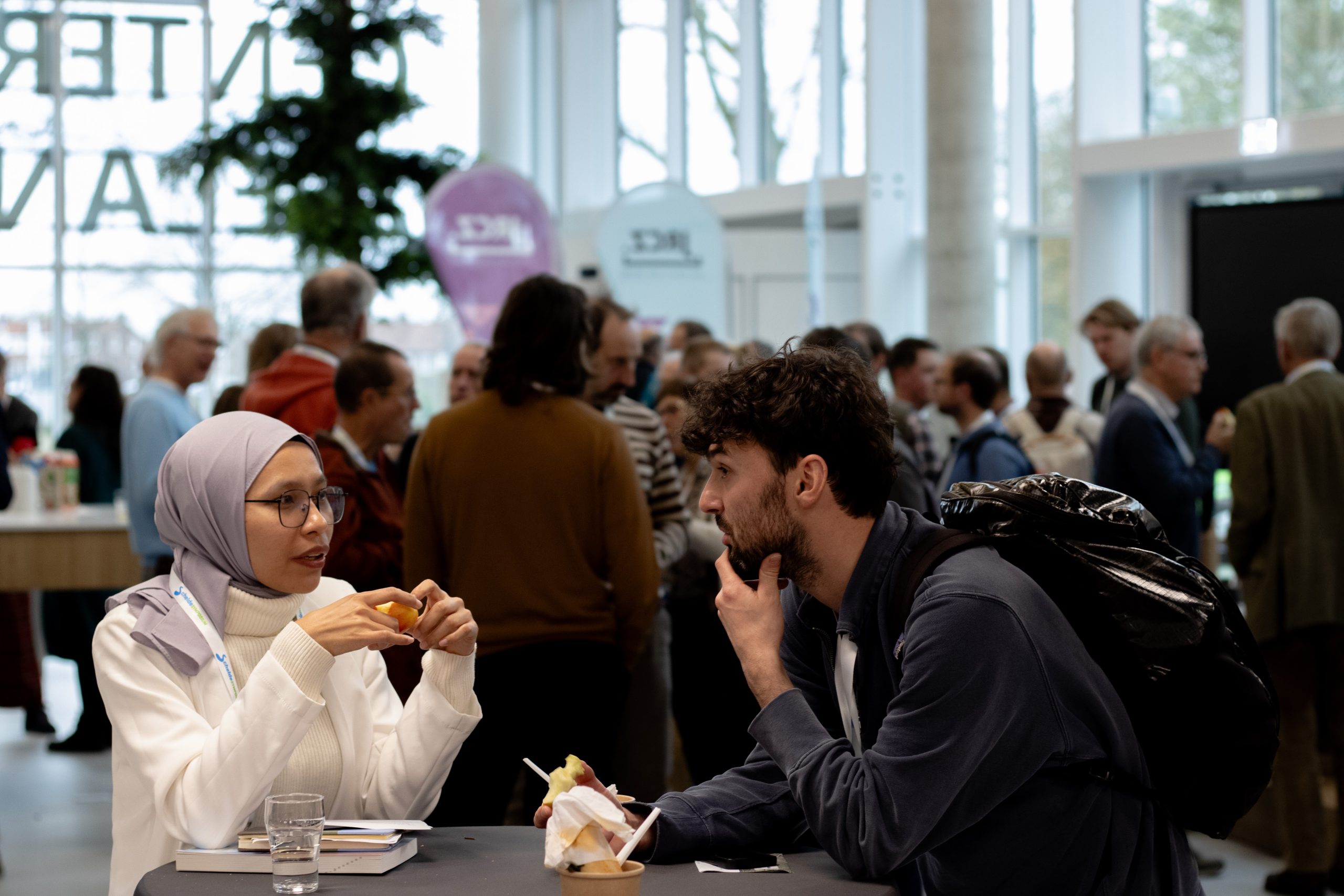 Twee deelnemers aan een statafel al pratend met een broodje in hun hand. De vrouw draagt een lila/grijze hoofddoek , een zwarte bril en een witte trui, de man heeft zwart kort haar en een donkerblauwe blouse. Hij draagt ook een zwarte rugzak.