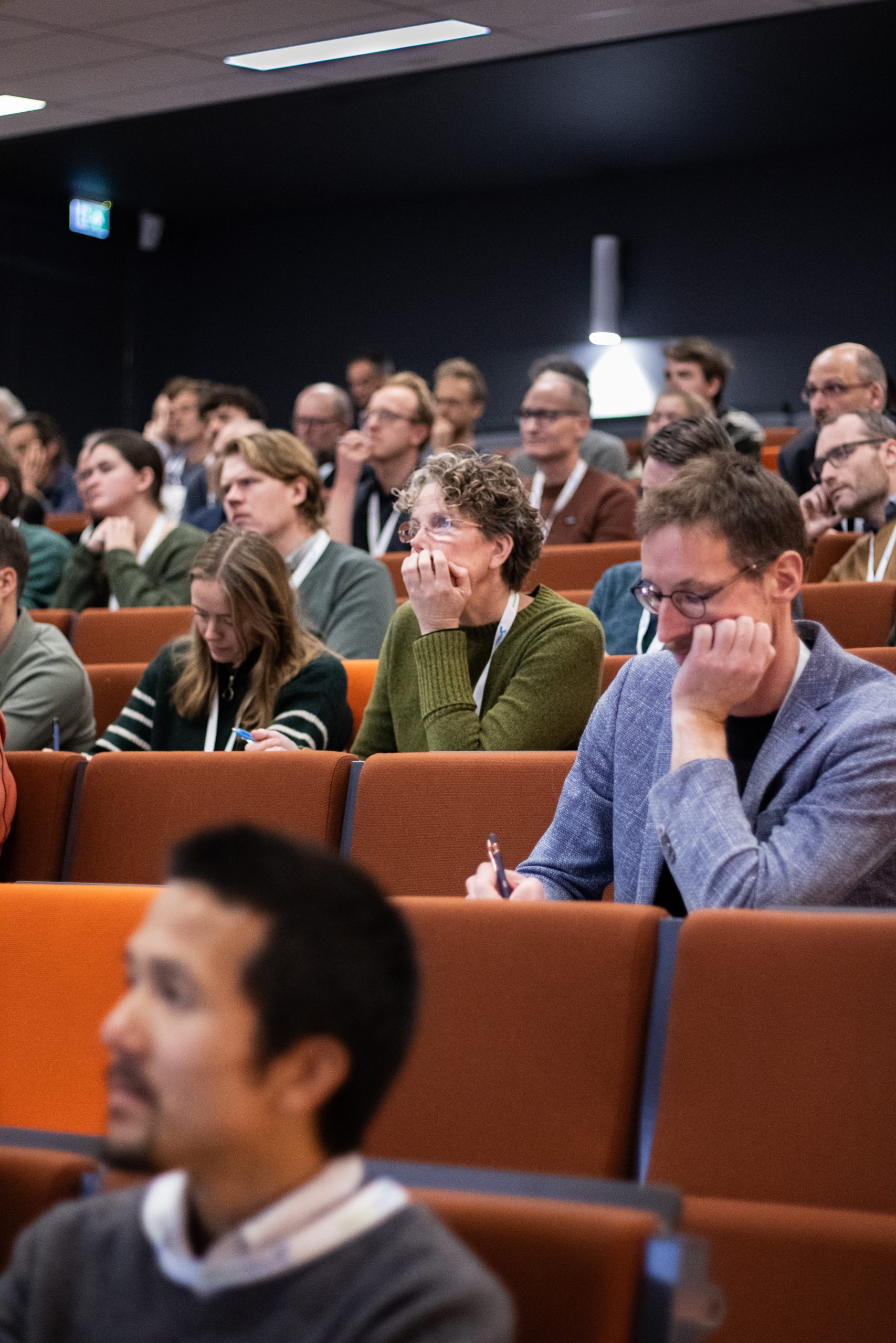 Close-up van het publiek Een blonde vrouw in de achtergrond maakt notities. De vrouw naast haar draagt een groene trui en leunt op haar hand terwijl ze geconcentreerd naar de presentatie kijkt. De man voor hen draagt een lichtblauwe blazer en een bril. Ook hij maakt notities.