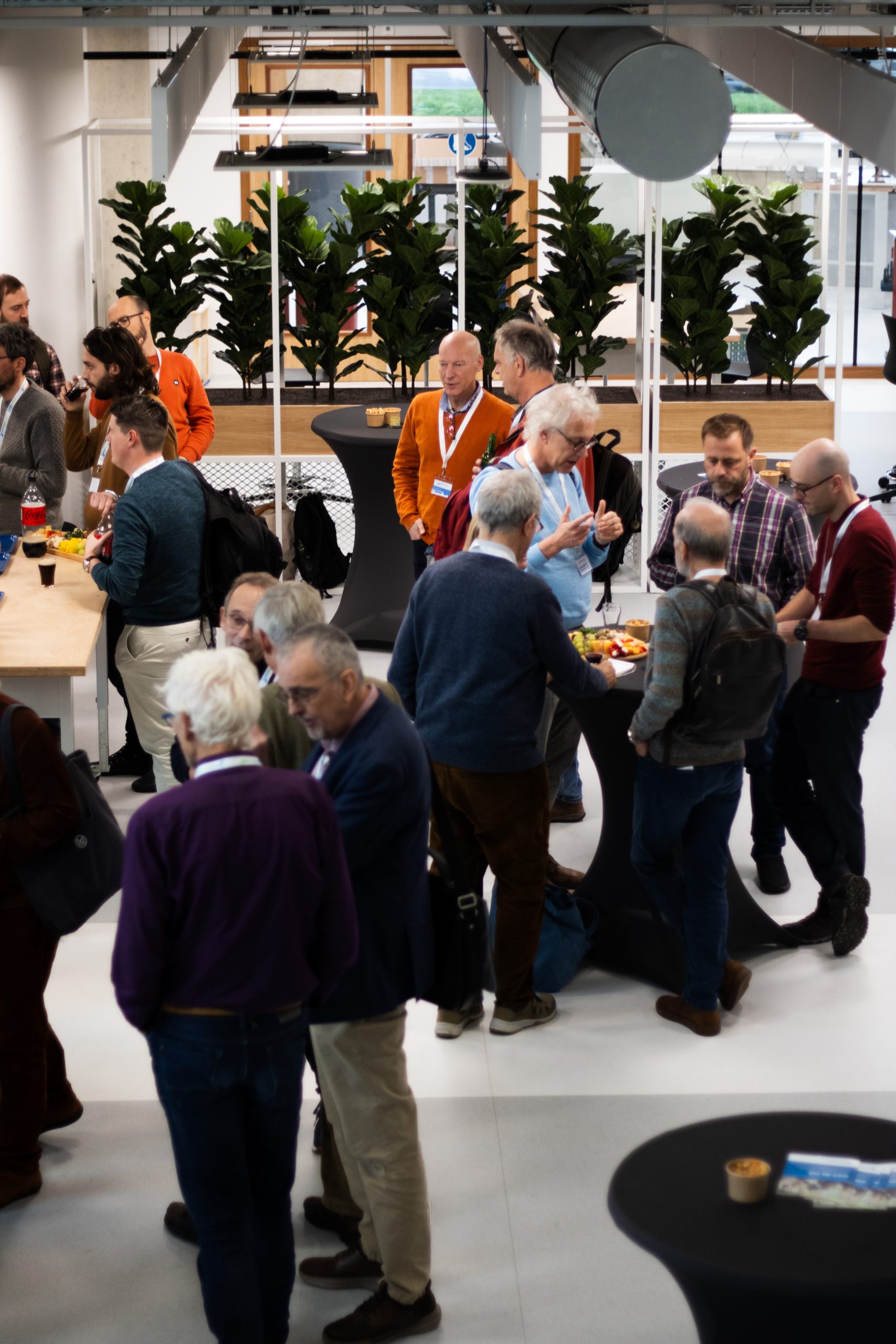 Foto van de naborrel van bovenaf. Mensen staan in groepjes bij diverse statafels met een drankje in hun hand.