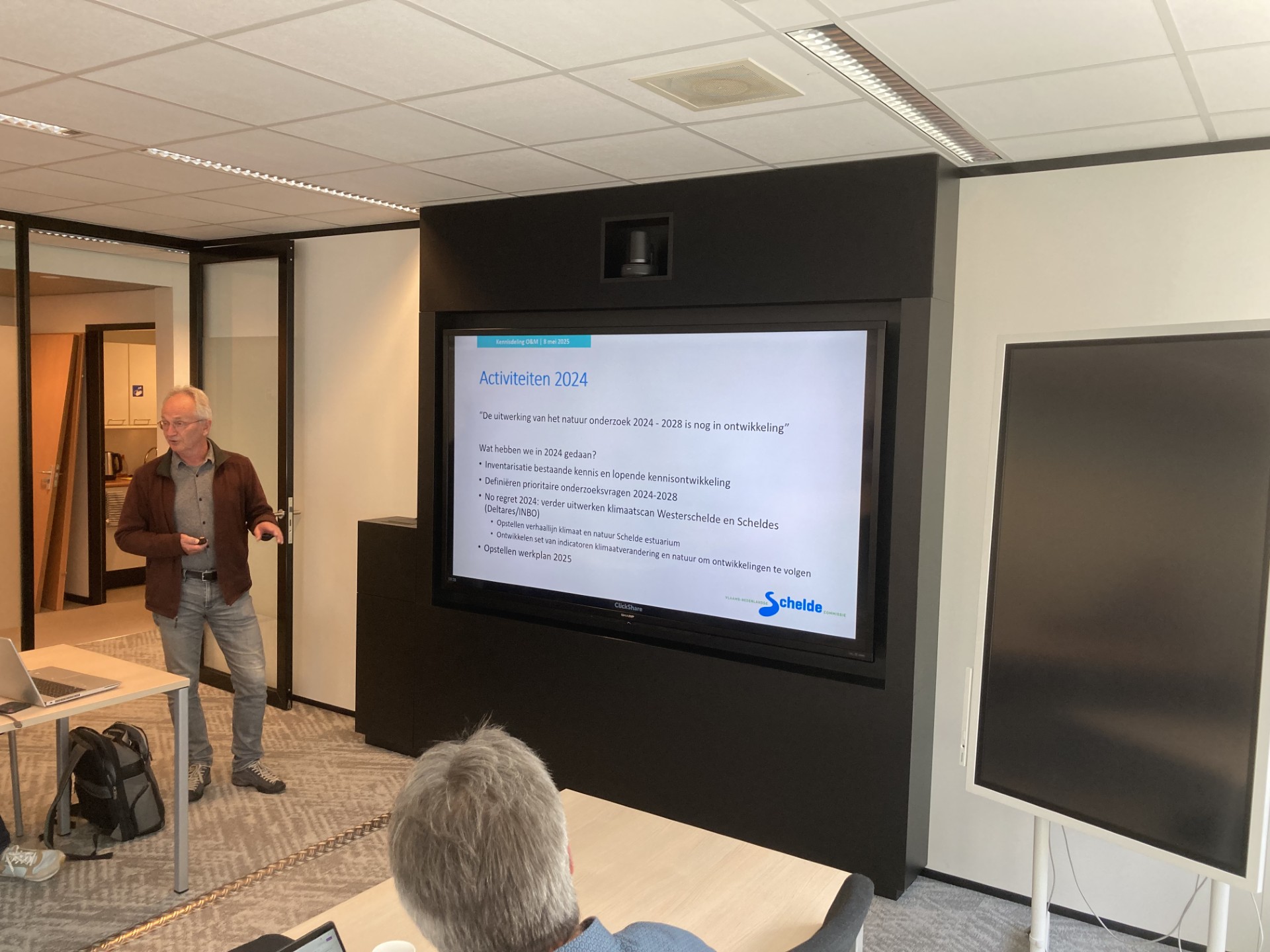 Een man staat bij een groot scherm en legt aan de toeschouwers in de zaal uit. Zij zitten aan een grote tafel.