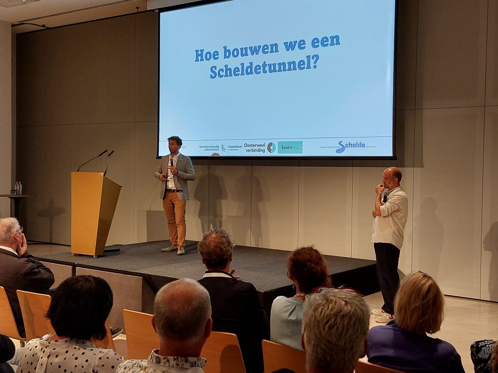 Presentatie tijdens de opening van het Scheldesymposium. Jonas Rabaey staat op het podium voor een beeldscherm. Hij spreekt namens Lantis, de opdrachtgever van de Oosterweelverbinding. Rechts van het podium staat Patrik Peeters van het Waterbouwkundig Laboratorium. Op het beeldscherm staat de vraag: "Hoe bouwen we een Scheldetunnel?"