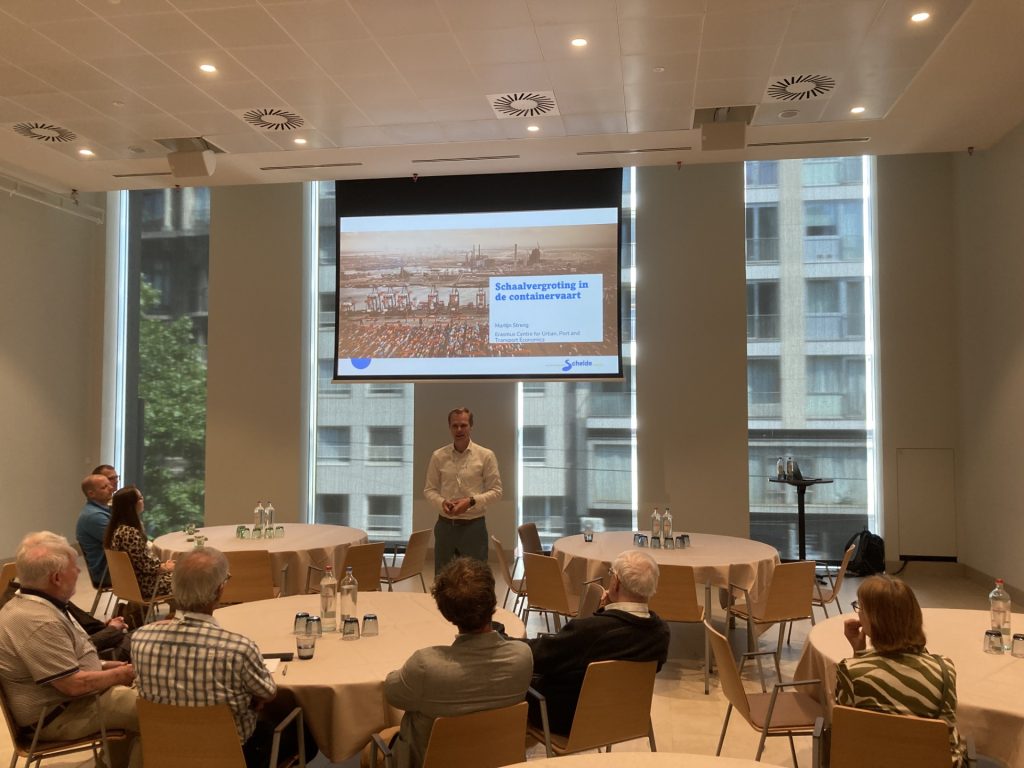 Presentatie schaalvergroting in de containervaart. Deelnemers aan het Scheldesymposium zitten aan ronde tafels. Spreker Martijn Streng staat voor het rolscherm. Te zien is de eerste dia met de titel van de break-outsessie.