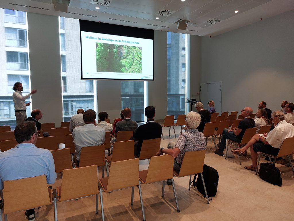 Presentatie Welzinge en Schorerpolder. De presentator in wit overhemd lacht en heft zijn arm op. Het rolscherm naast hem laat een luchtfoto te zien van een groene omgeving. De tekst op het scherm is "Welkom in Welzinge en de Schorerpolder".