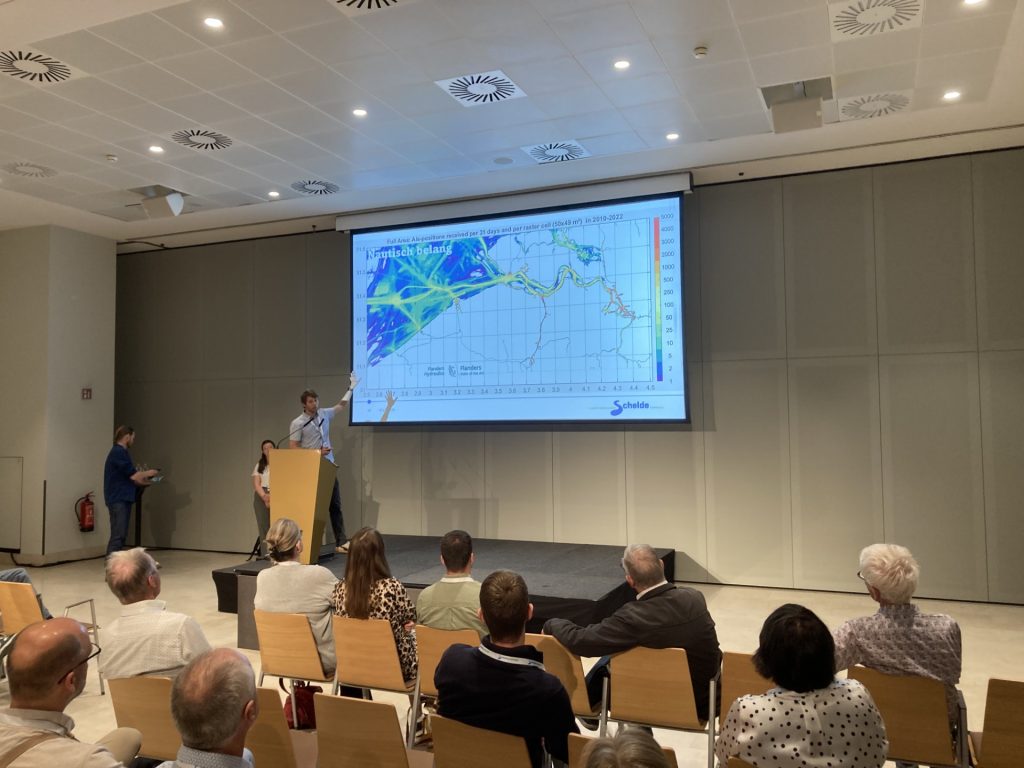 Presentatie Schaar van Valkenisse. Drie presentators staan bij een rolscherm. Het publiek zit op stoelen. Op het rolscherm staat de tekst "Nautisch belang". Je ziet een kaart van het Schelde-estuarium in de periode 2010-2022. Uit de kaart blijkt dat de Westerschelde flink gebruik wordt door de scheepvaart.